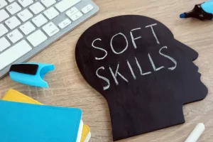 Silhouette-shaped chalkboard on a desk displays the words "SOFT SKILLS" near a keyboard, notebooks, and a marker.