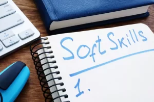 Open notebook with "Soft Skills" written on a page, placed on a wooden desk next to a keyboard, a blue mouse, and a closed blue planner.