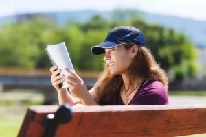 A person wearing a cap and a maroon shirt is sitting on a wooden bench outdoors, smiling while reading a book.