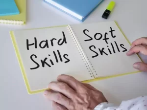 Person's hands hold an open notebook labeled "Hard Skills" on the left page, "Soft Skills" on the right. A highlighter and notebooks are visible on the table.
