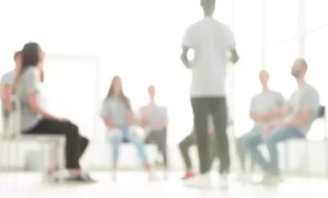 A group of people sits in a circle while one person stands, leading a discussion in a brightly lit room.