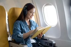 A person wearing headphones reads a book while sitting in an airplane seat next to a window.