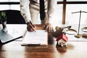 A person writes on a clipboard at a desk with a laptop, gavel, scales of justice, and a small house model.