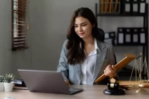 A woman in a gray blazer holds a folder and looks at a laptop on a desk, with a gavel and scales nearby.