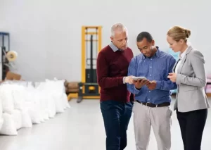 Three people in a warehouse reviewing information on a tablet, with machinery and bags in the background.