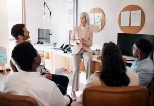 A woman sits on a desk, speaking to four seated people in an office setting with a screen, lamp, and corkboards.