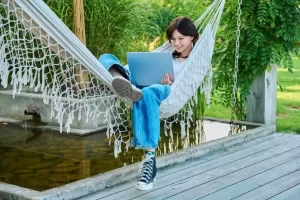 A person sitting in a hammock using a laptop outdoors, surrounded by greenery and a small pond.