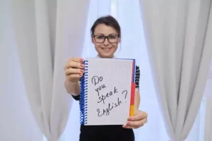 Person holding a notebook with the handwritten question "Do you speak English?" in front of a curtain backdrop.
