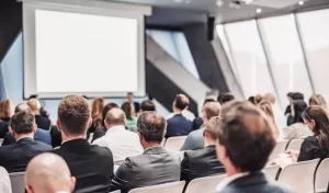 Audience attending a presentation in a conference room with a large screen at the front.