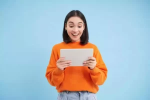 A woman in an orange sweater looks surprised while holding a tablet against a blue background.