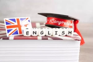 Graduation cap and small UK flag on stacked paper with letter cubes spelling "ENGLISH.