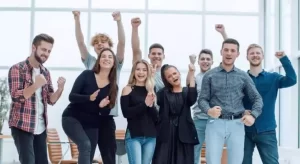 A group of eight people standing indoors, smiling and cheering, with modern office chairs and a large window in the background.