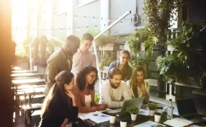 A diverse group of people gathered around a laptop at a table in an outdoor setting with greenery and sunlight.