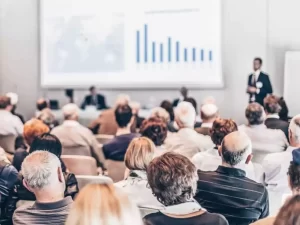 Audience seated in a conference room listens to a presentation with a visible bar chart on a large screen.