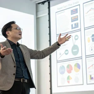 Presentational Skills & Storytelling Techniques - Start embracing storytelling to engage, influence, and bring your ideas to life 285 A man in a suit gestures while presenting data charts and graphs on a projector screen to an audience in a meeting or classroom setting.