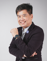 A person with short dark hair wearing a dark blazer and light shirt is smiling and posing with their right arm crossed over their chest against a plain light background.