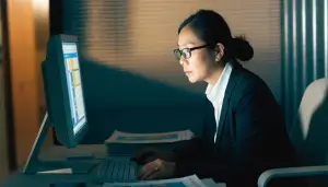 A professional woman wearing glasses sits at a desk using her AI problem-solving skills on a computer in a dimly lit office.