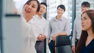 A group of people in an office, with a woman smiling and pointing at a whiteboard while others look on attentively.