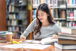 A person sits at a library table, writing in a notebook. A stack of books, a highlighter, and a coffee cup are nearby. Shelves filled with books are visible in the background.