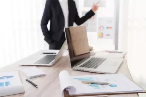 A business person points at charts on a wall. Two open laptops, documents, a pen, and a notebook are on a table.