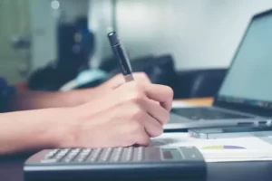 A person writes in a notebook beside a calculator and an open laptop on a desk.