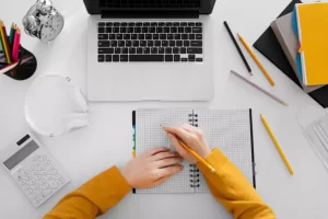 Person writing in a notebook at a desk with a laptop, calculator, headphones, and various stationery items.