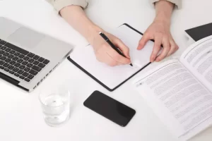 Person writing in a notebook at a desk with a laptop, open book, smartphone, and a glass of water.