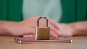 A padlock sits on top of a smartphone, symbolizing security. The background is blurred, and a person's folded hands are partially visible.