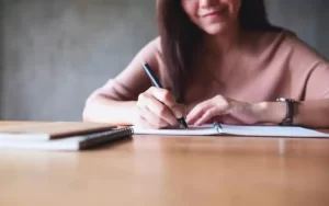 A person sits at a wooden table, writing in a notebook with a pen. Another notebook lies nearby.