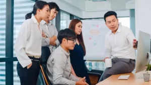 A group of people in a modern office are gathered around a computer, discussing something on the screen. One person is sitting on a desk, holding a mug, and pointing at the monitor.