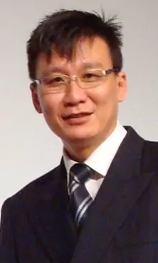 A person with short dark hair, wearing glasses and a suit with a striped tie, smiling at the camera.