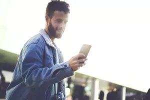 A man in a denim jacket smiles while looking at his smartphone outdoors.