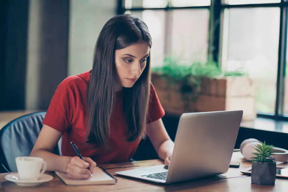 A woman in a red shirt is focused on her laptop while taking notes at a wooden table. A cup and plant are nearby.