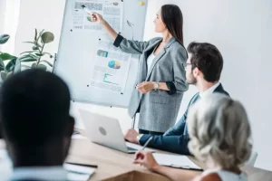 A woman points to a chart on a whiteboard during a meeting, while three colleagues sit around a table with laptops.