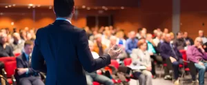 A person in a suit speaks to a seated audience in a conference room.