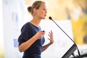 A woman is speaking into a microphone at a podium, gesturing with one hand.