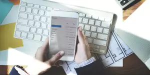 Person using a smartphone displaying an email inbox, sitting at a desk with a keyboard and documents.