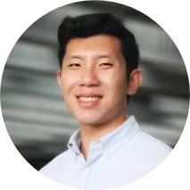 A person with short dark hair wearing a dark blazer and light shirt is smiling and posing with their right arm crossed over their chest against a plain light background.