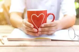 A person holds a red mug with a heart design, placed on an open notebook on a wooden table, with glasses and a pen nearby.
