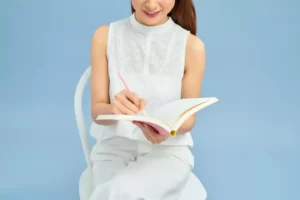 A person sitting on a chair, writing in a notebook with a pink pencil, wearing a sleeveless white top and pants, against a light blue background.