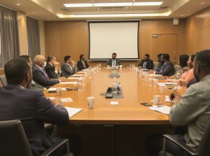 A group of people in business attire sit around a long conference table in a meeting room with papers, cups, and a projector screen visible.