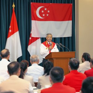 A man speaks at a lectern in front of a Singapore flag, addressing an audience seated in rows. Two smaller Singapore flags stand on either side.