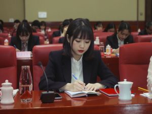 A woman in formal attire writes in a notebook at a conference table, surrounded by others attending the meeting.