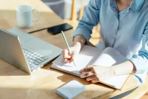Person writing in a notebook at a desk with a laptop, smartphone, and coffee mug.