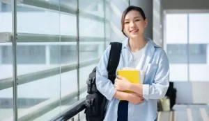 A person smiles while holding notebooks and a backpack, standing in a well-lit hallway with large windows.