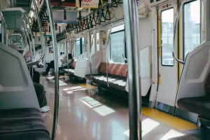 An empty train car with rows of seats on either side, metal poles, and large windows allowing sunlight inside.