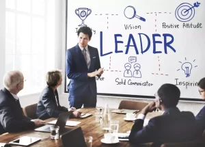 A man in a suit presents a leadership concept on a whiteboard to a group of seated professionals in a conference room.