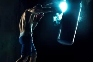 A person wearing boxing gloves practices with a punching bag, illuminated by a dramatic blue light.