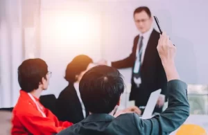 A person raises a pen in a meeting while another stands and speaks, pointing at a board. Two others are seated, listening attentively.