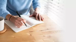 Person writing in an open notebook with a pen at a wooden table, next to a white coffee mug.